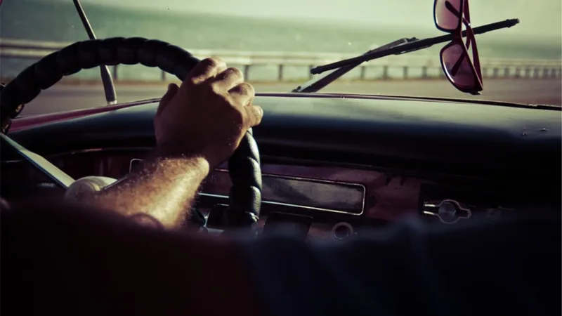 Mains d'un conducteur sur le volant d'une voiture ancienne, lunettes de soleil accrochées au pare-soleil.