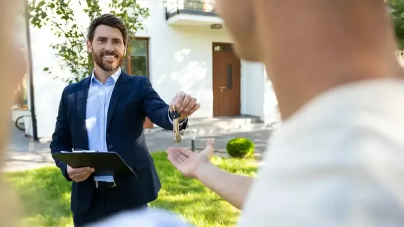 Un agent immobilier en costume remet les clés d'une maison à un acheteur, devant la maison.