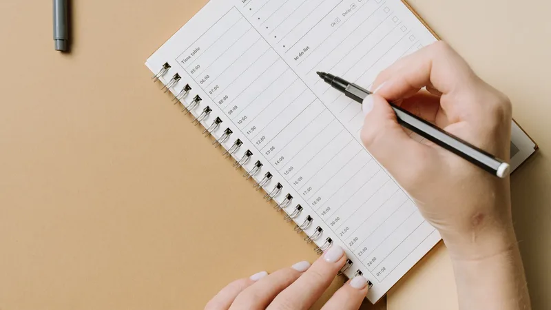Un agenda à spirale vu de dessus sur un bureau beige, une main y écrit au stylo à côté d'un crayon : planifier et compter les jours de la semaine.