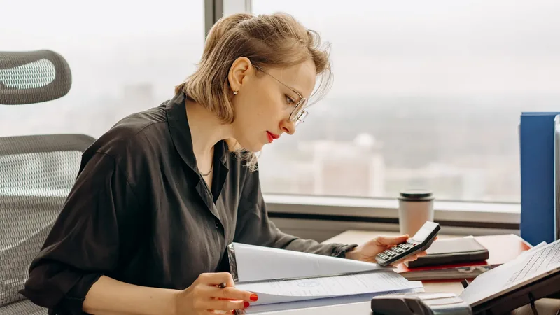 Une salariée à son bureau relit un dossier de documents, calculatrice à la main, devant une grande fenêtre : la vérification ligne par ligne d'un bulletin de paie.