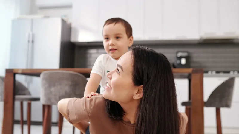 Une mère souriante porte son jeune fils sur son dos dans une cuisine lumineuse : les besoins quotidiens de l'enfant que la pension alimentaire couvre.