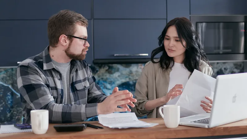 Un couple assis à la table de sa cuisine discute des papiers étalés devant lui, ordinateur portable ouvert : arbitrer entre remboursement du crédit et épargne.